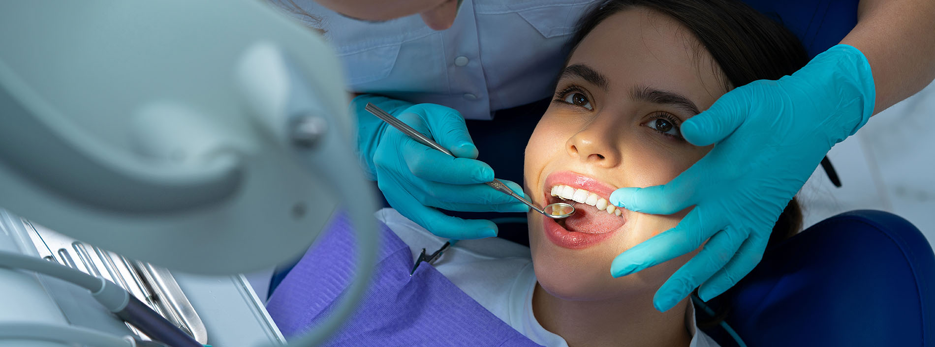 A woman receiving dental treatment with an open mouth, while a dentist is examining her teeth with a magnifying glass.