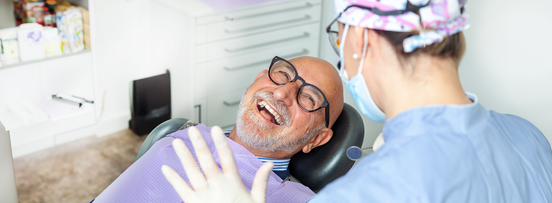 A man in glasses is seated in an examination chair with his mouth open, receiving dental care from a person wearing a surgical mask and blue scrubs, set against the backdrop of a dental office interior.