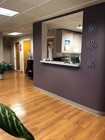 The image shows an interior view of a waiting room with a reception desk, a countertop, a framed picture on the wall, a potted plant, and a sign with a phone number.