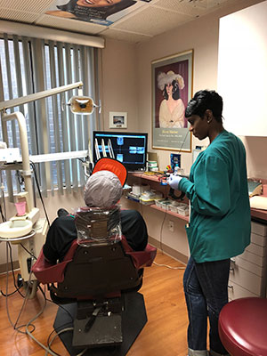 A dental office with a dentist seated at a chair, a dental hygienist standing next to him, and various equipment visible, including a computer screen displaying an X-ray image.