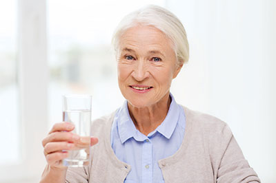 The image shows an older woman holding a clear glass filled with water while standing indoors, smiling slightly towards the camera.