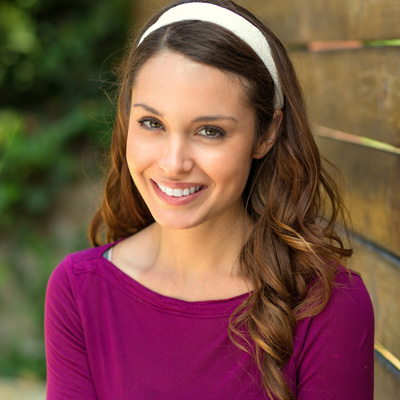 A young woman with long hair, wearing a purple top and a headband, smiling at the camera against a wooden fence background.