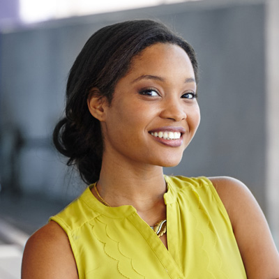 A smiling woman with dark hair wearing a yellow top and standing against a wall.