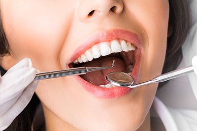 A woman with open mouth receiving dental care while wearing a white lab coat, holding dental instruments.