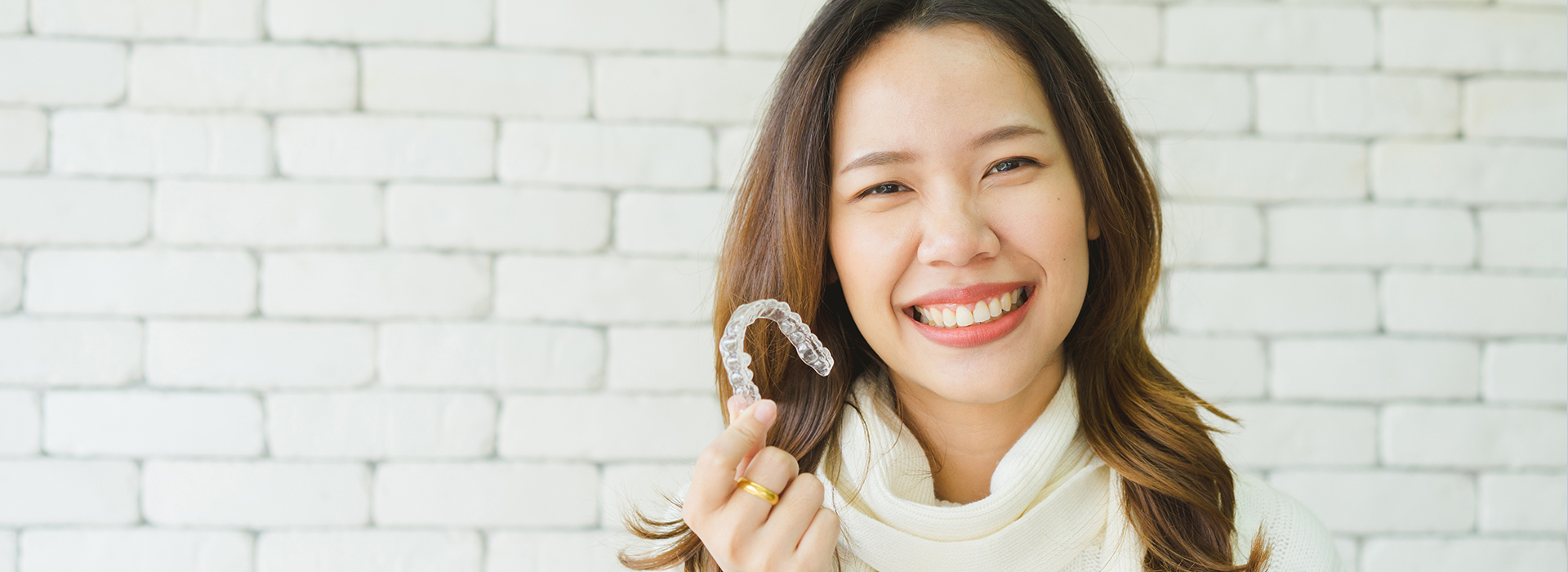 A woman with a smile, wearing a ring, stands against a brick wall.