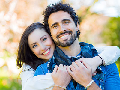 The image depicts a man and woman embracing each other with smiles on their faces, set against an outdoor background.