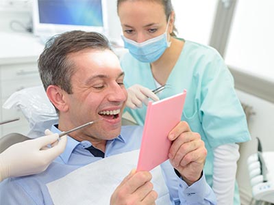 A man sitting in a dental chair, holding up a pink card with a surprised expression, while a dentist looks on behind him.