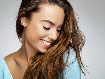 The image features a young woman with long brown hair smiling gently while looking downwards, with her head slightly tilted towards the viewer. She appears to be wearing a light blue top.