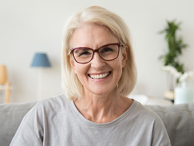 The image shows a woman with short blonde hair, wearing glasses, smiling at the camera, set against a blurred indoor background.