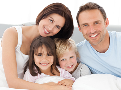 A family of four poses together on a bed with smiles, including an adult couple and two children.