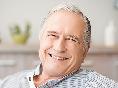 The image shows an elderly man with gray hair, wearing glasses and a blue shirt, smiling and looking directly at the camera, sitting comfortably in a chair.