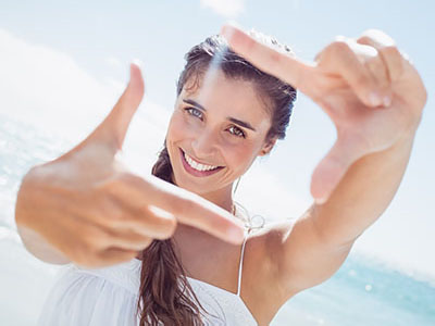 A young woman with long hair smiles at the camera while holding up her index finger, set against a bright beach backdrop.